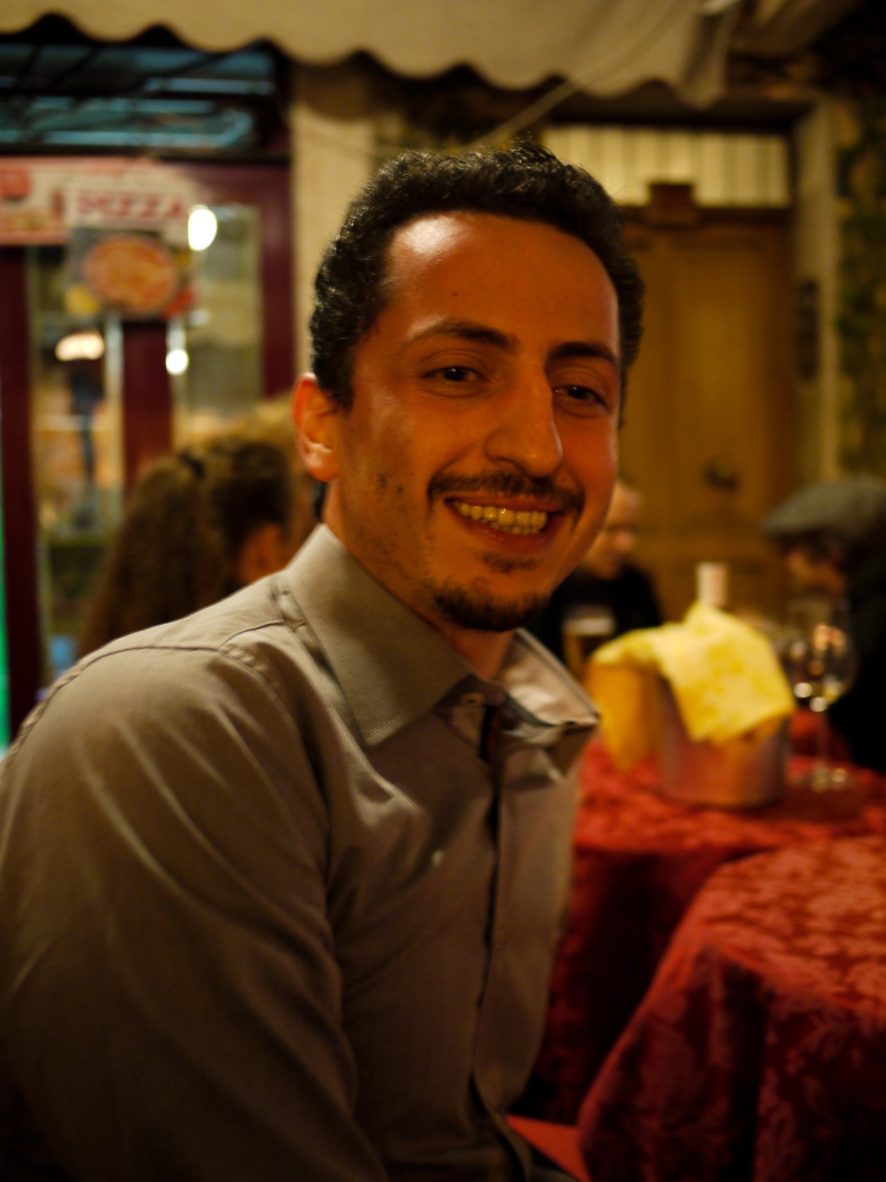 A man in a gray shirt smiles while sitting at a restaurant table with drinks and food in Rome.
