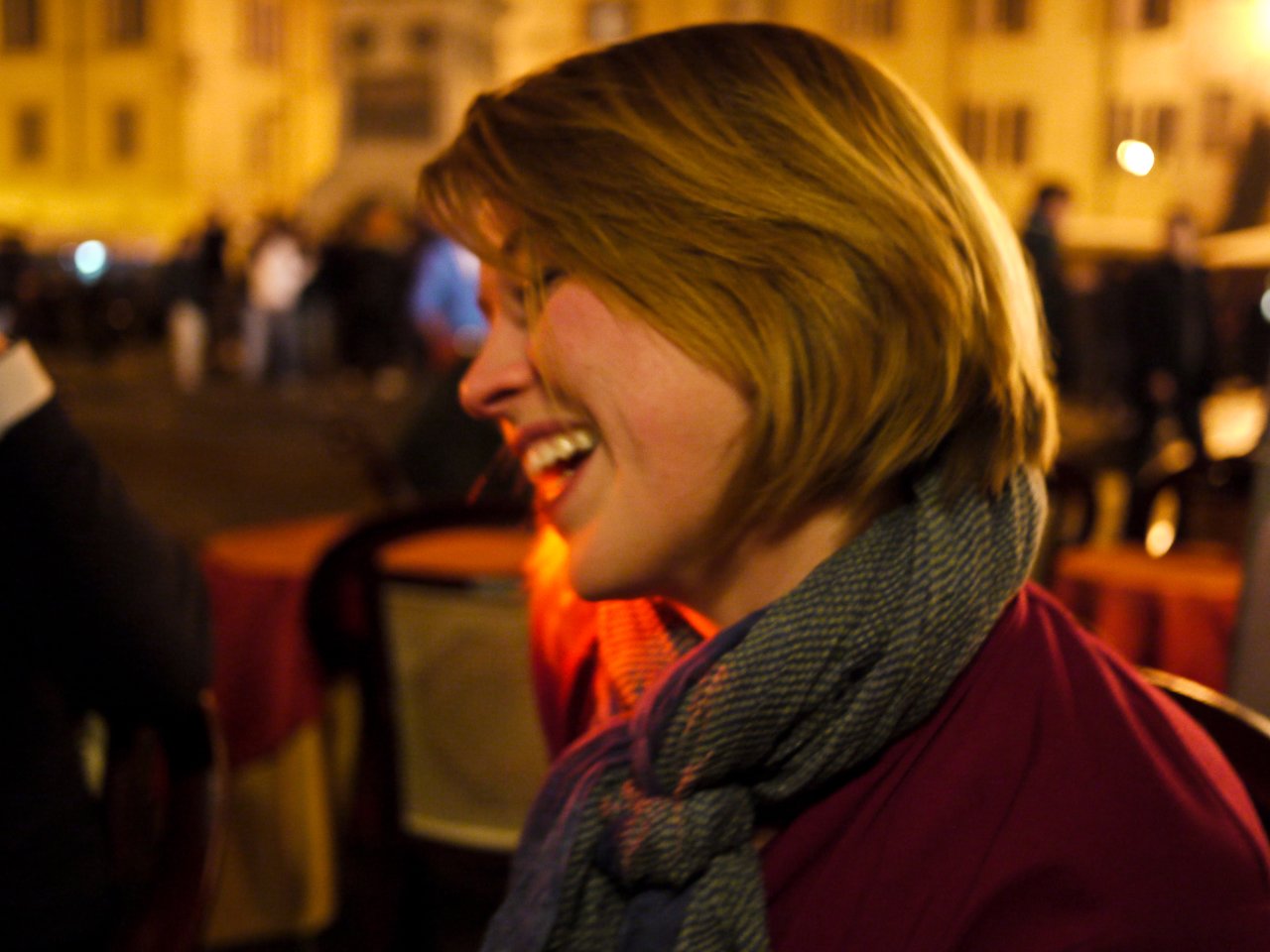 A woman with short hair and a scarf laughs while sitting at an outdoor café in Rome at night.
