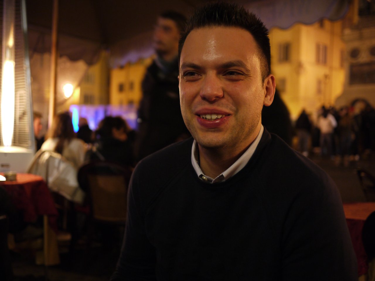 A man in a dark sweater smiles while sitting at an outdoor restaurant in a busy nighttime setting.