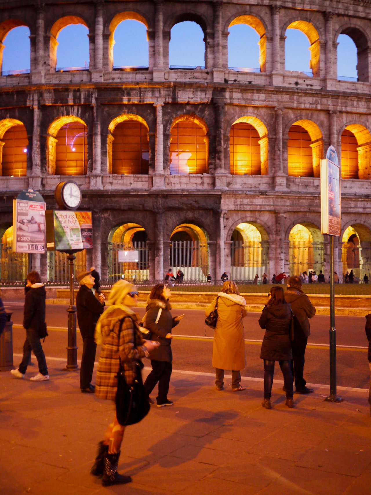 People stand near the illuminated Colosseum in Rome during the evening, some taking photos and others observing the scene.