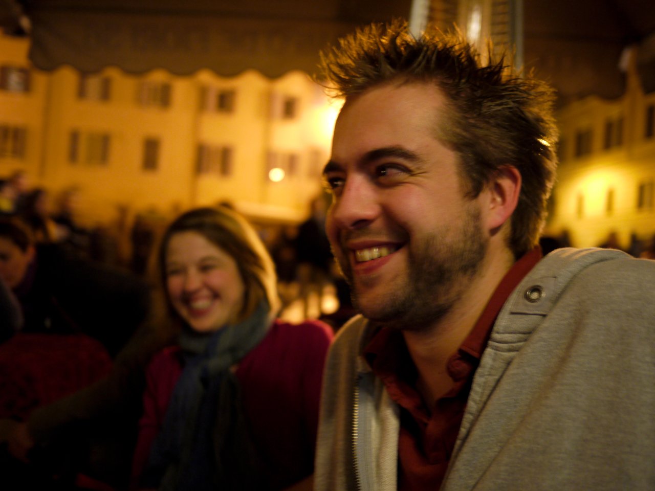 A man in a gray hoodie and a woman in a scarf smile while sitting outdoors at night.