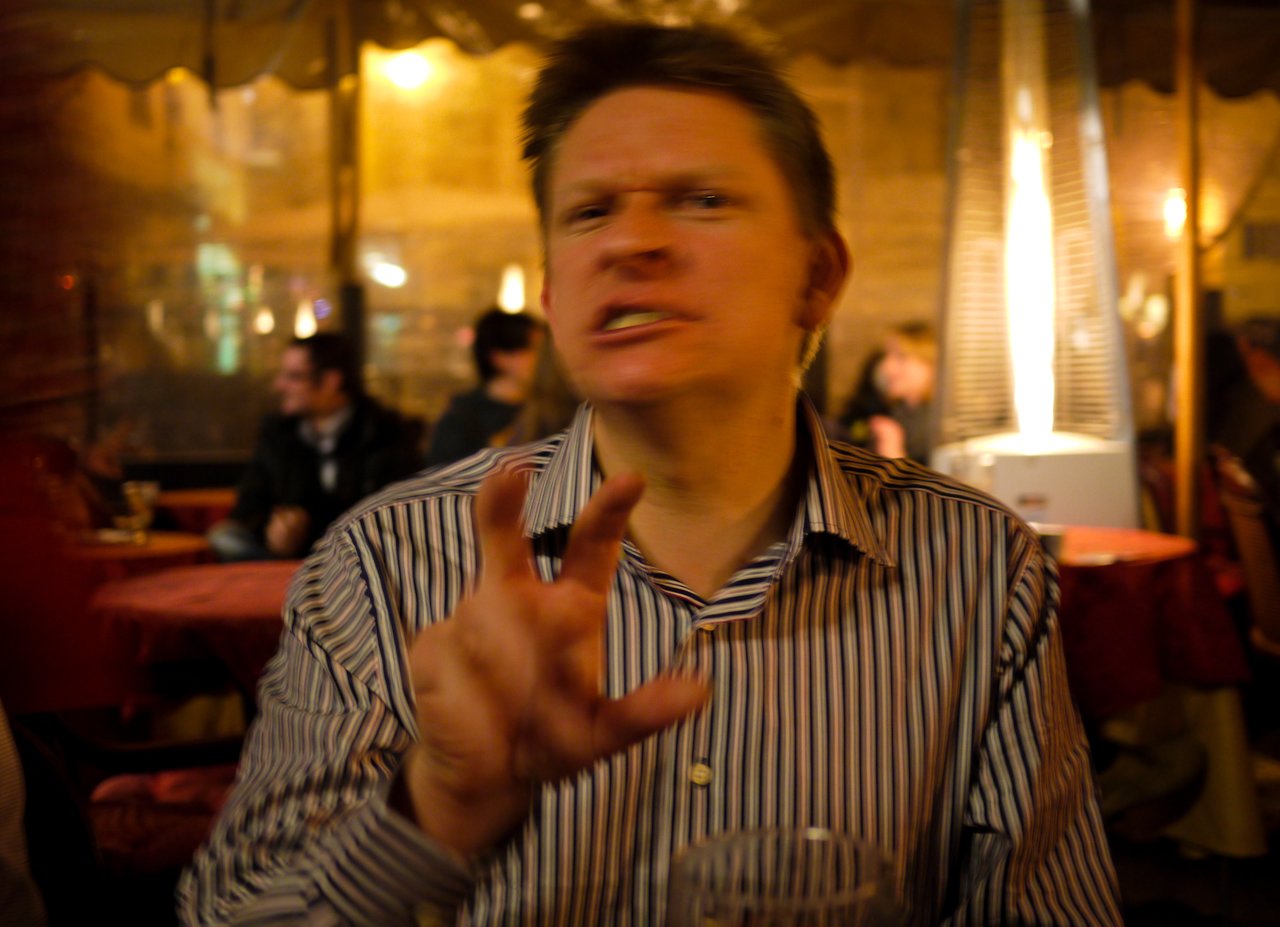 A man in a striped shirt gestures while speaking at a restaurant with warm lighting.