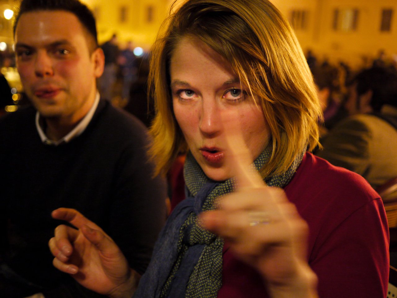A woman in a scarf gestures with her hands while speaking, with a man sitting nearby in a crowded setting.