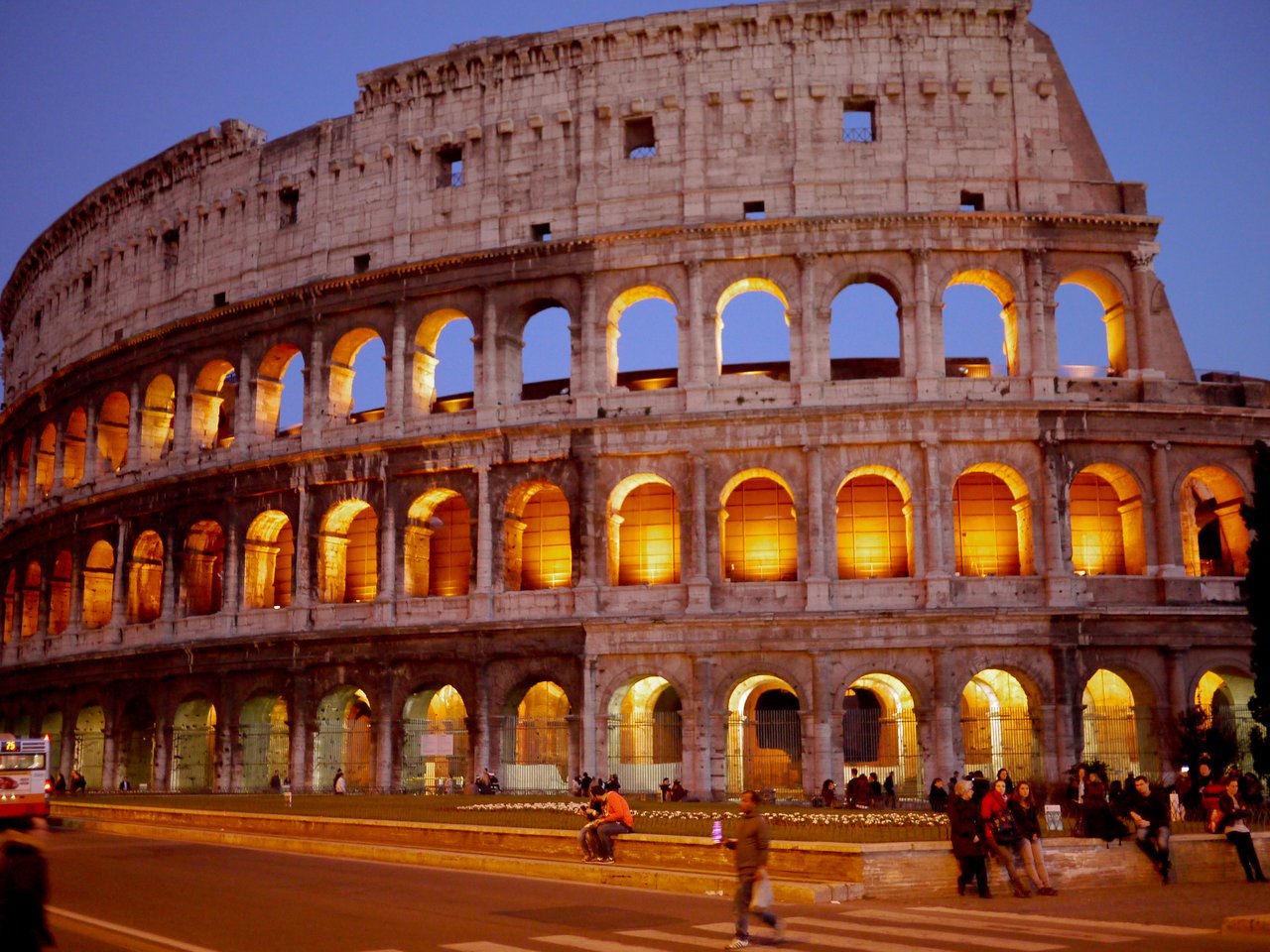 The Colosseum in Rome is illuminated at dusk, with people walking and sitting nearby.