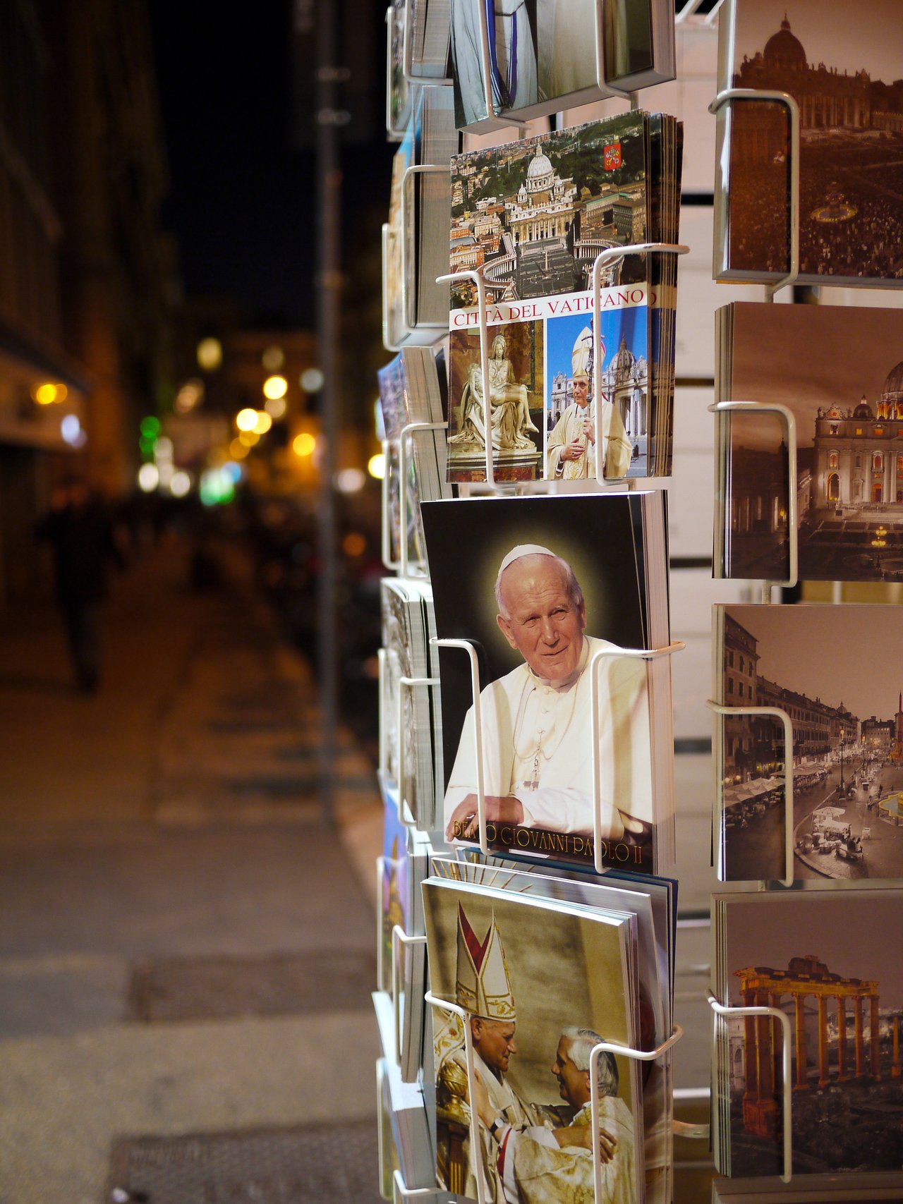 A postcard stand displays images of Rome and the Vatican, including a portrait of a pope, on a street.