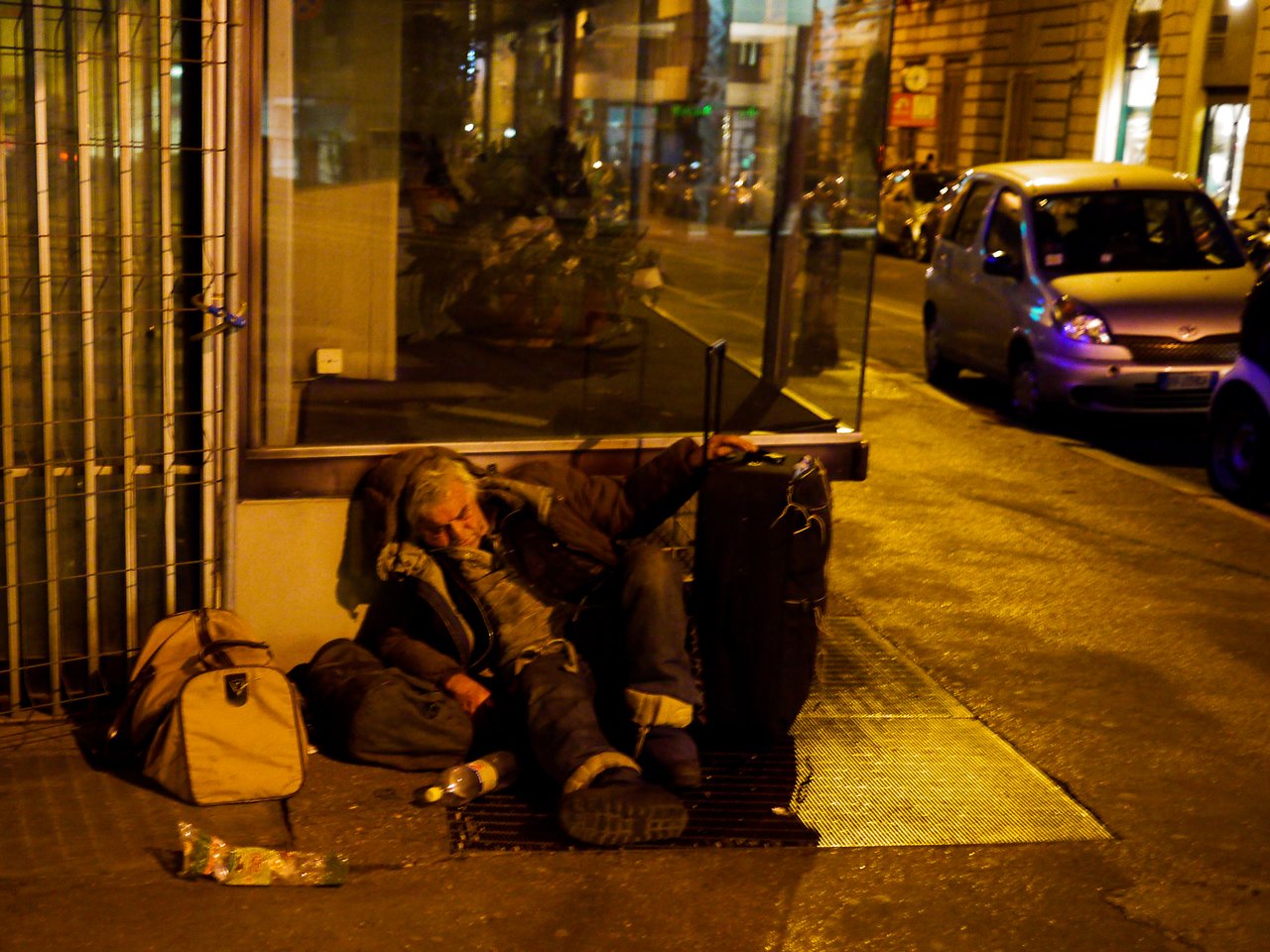 A person is sleeping on the sidewalk, leaning against luggage, with bags and bottles nearby at night.