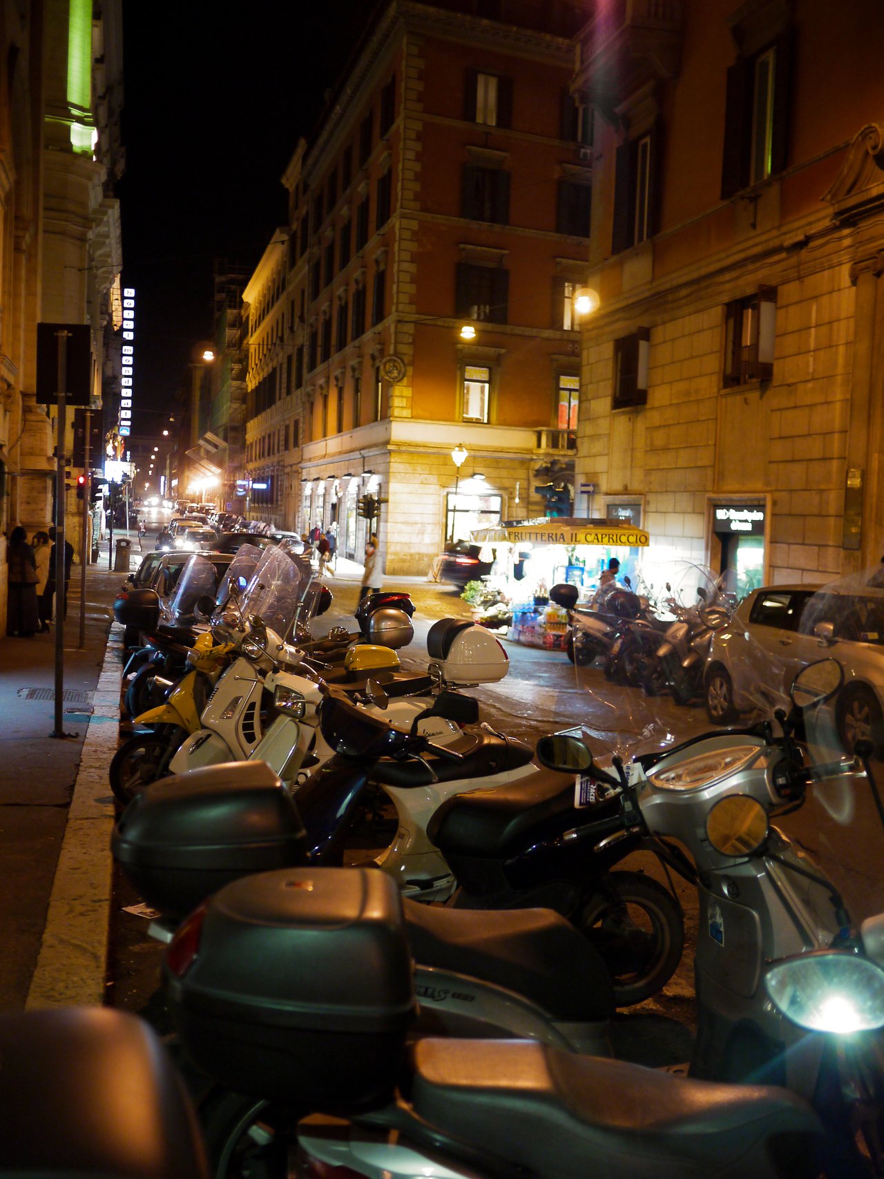 Rows of parked scooters line a busy street at night, with buildings and storefronts illuminated in the background.