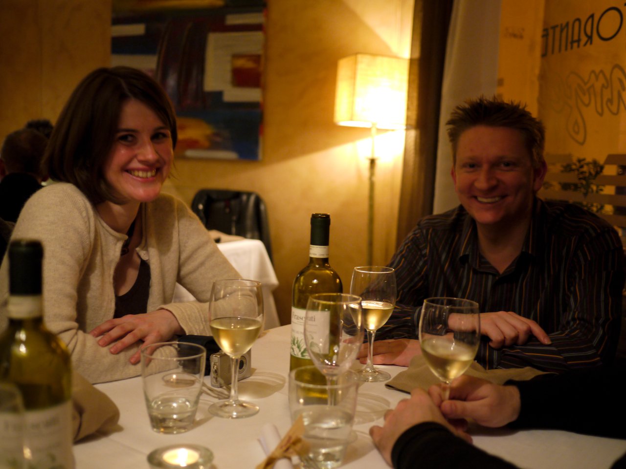 Two people smiling at a restaurant table with wine glasses and bottles, engaged in conversation with others.