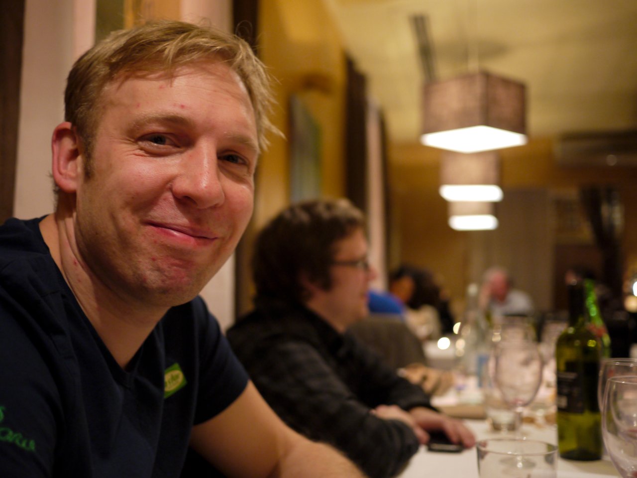 A man in a dark shirt smiles at the camera while sitting at a restaurant table with others.