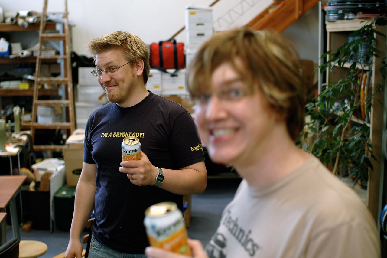 Two people in a workspace holding canned drinks, one smiling in the foreground and the other smirking in the background.