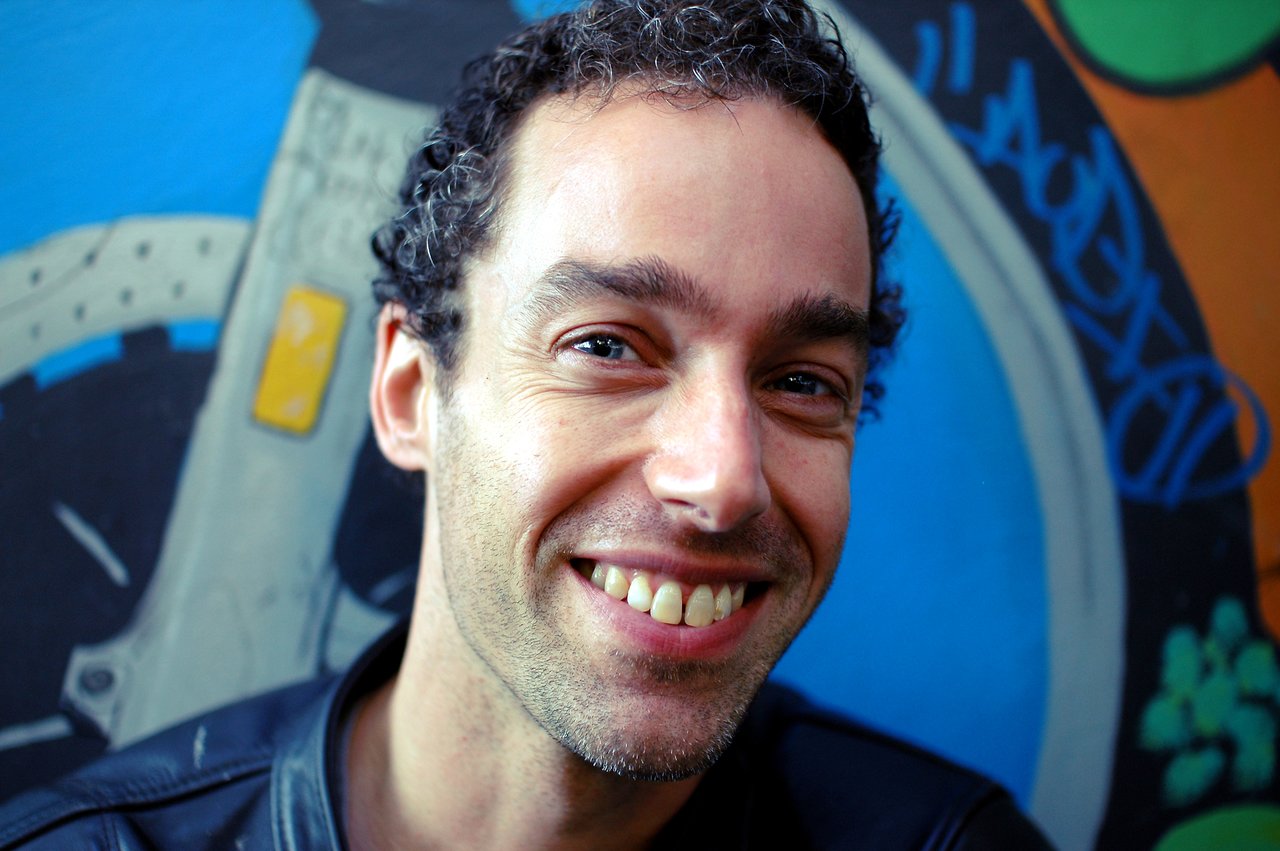 A man with short curly hair smiles at the camera, standing in front of a colorful graffiti wall.