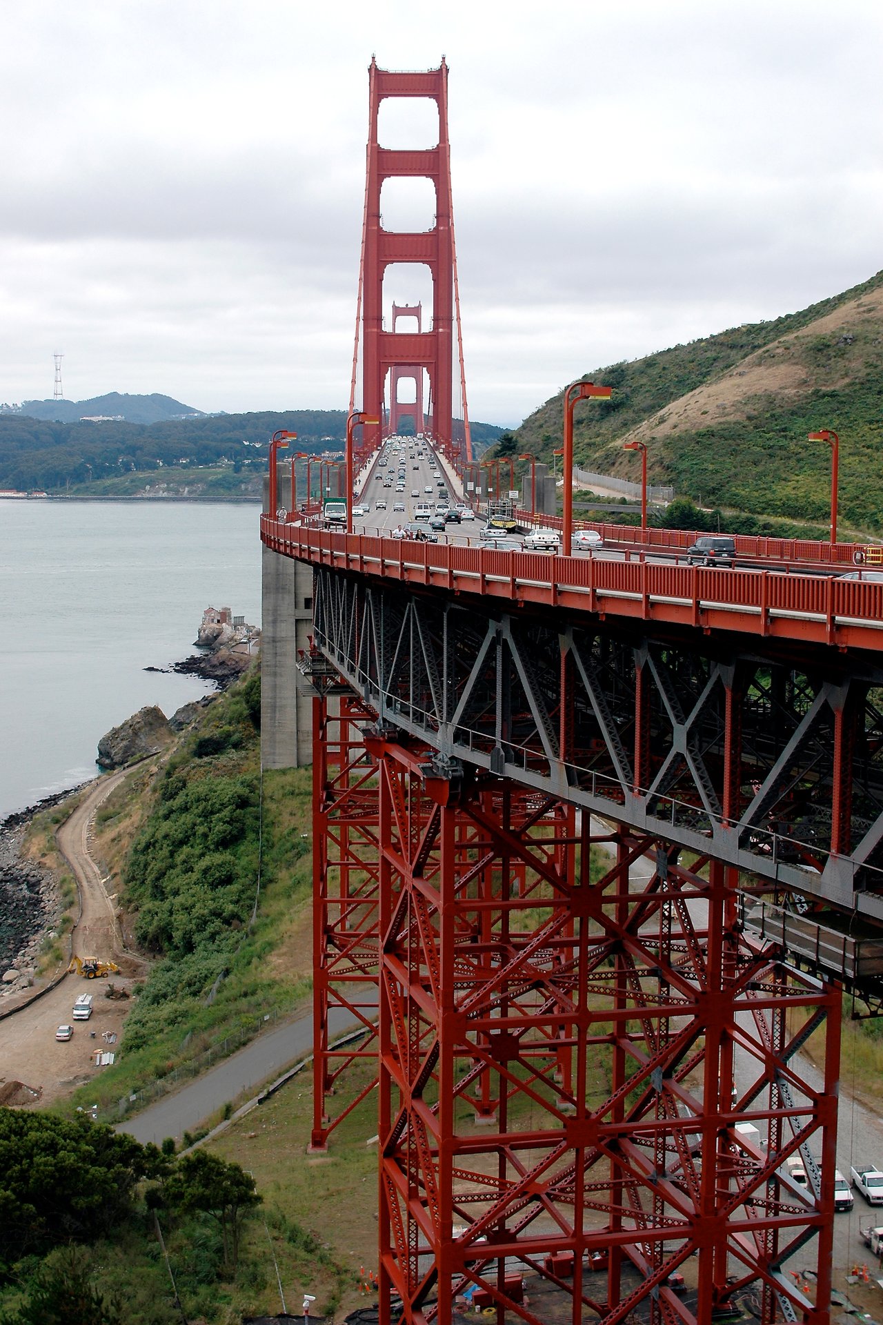 Cars drive across the Golden Gate Bridge, supported by red steel beams, under an overcast sky.