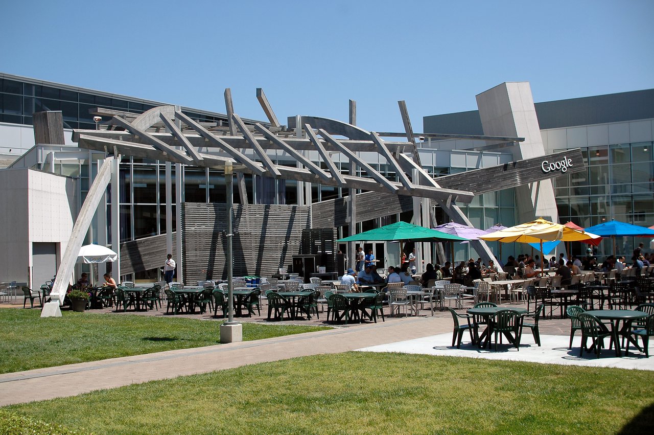 People sit at outdoor tables under colorful umbrellas on a patio at a Google office building.