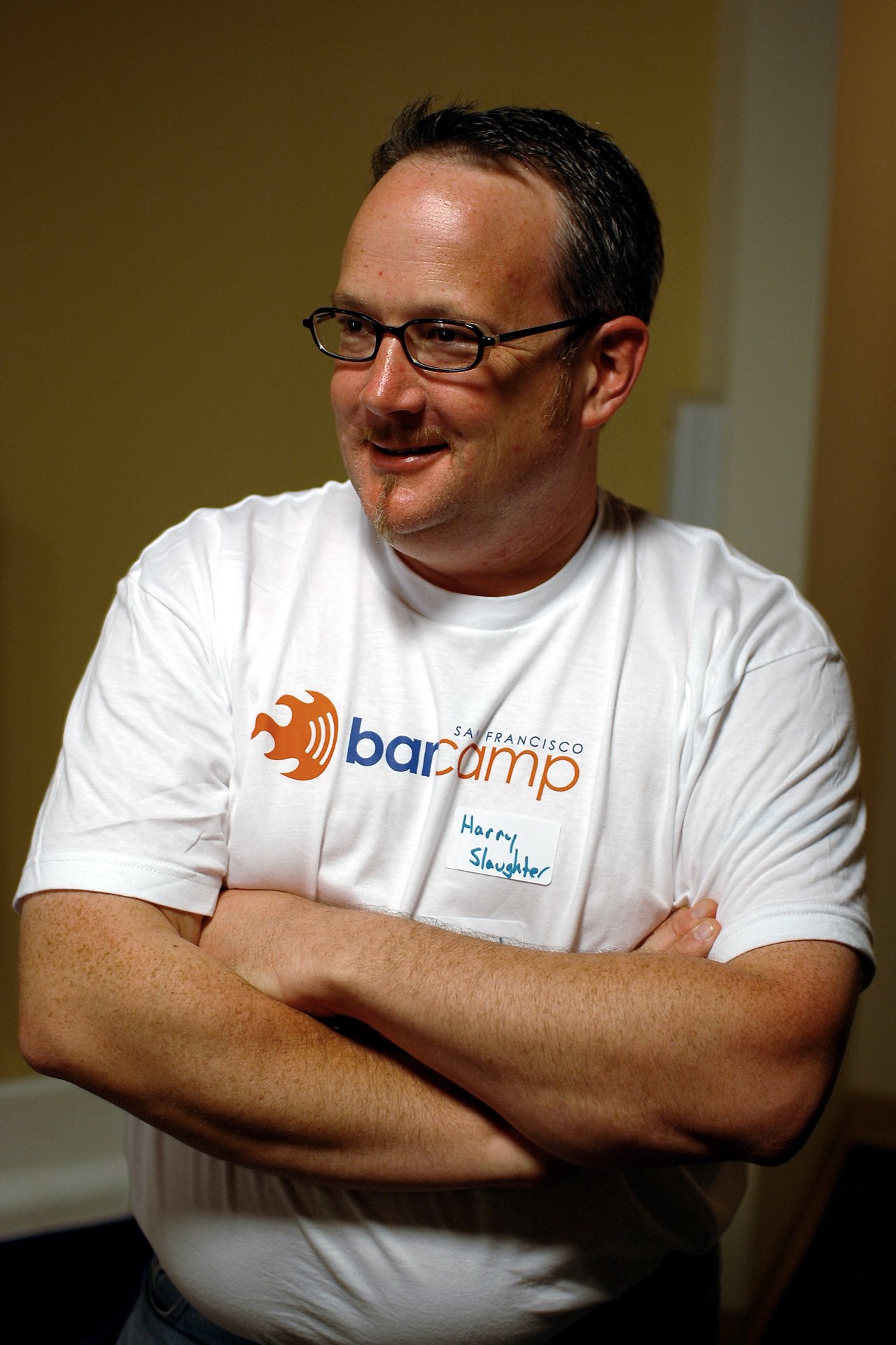 A man wearing a BarCamp San Francisco t-shirt and name tag stands with arms crossed, smiling.