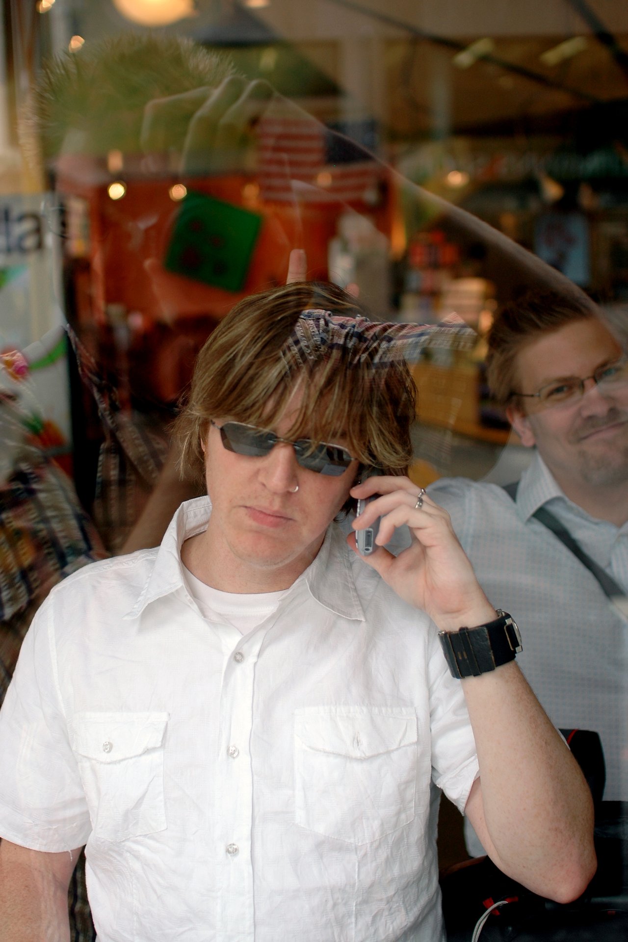 A man in sunglasses and a white shirt talks on his phone, while another man smiles behind a glass window.