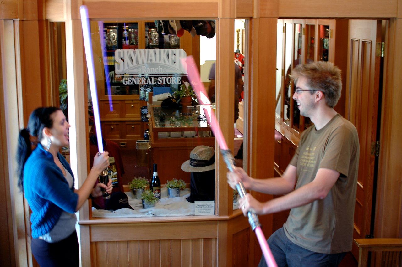 Two people smiling and playfully dueling with toy lightsabers inside a store with a "Skywalker Ranch General Store" sign.