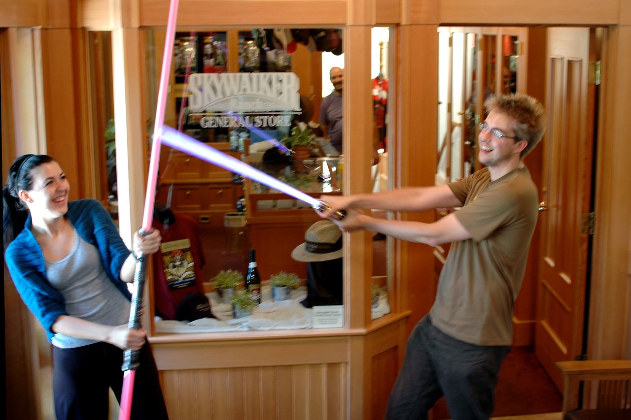 Two people smiling and playfully dueling with toy lightsabers inside a store with a "Skywalker Ranch General Store" sign.