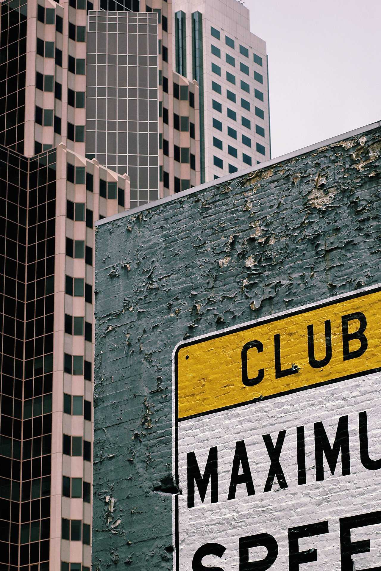 A weathered wall with a partially visible "Maximum Speed" sign, set against tall modern buildings in the background.