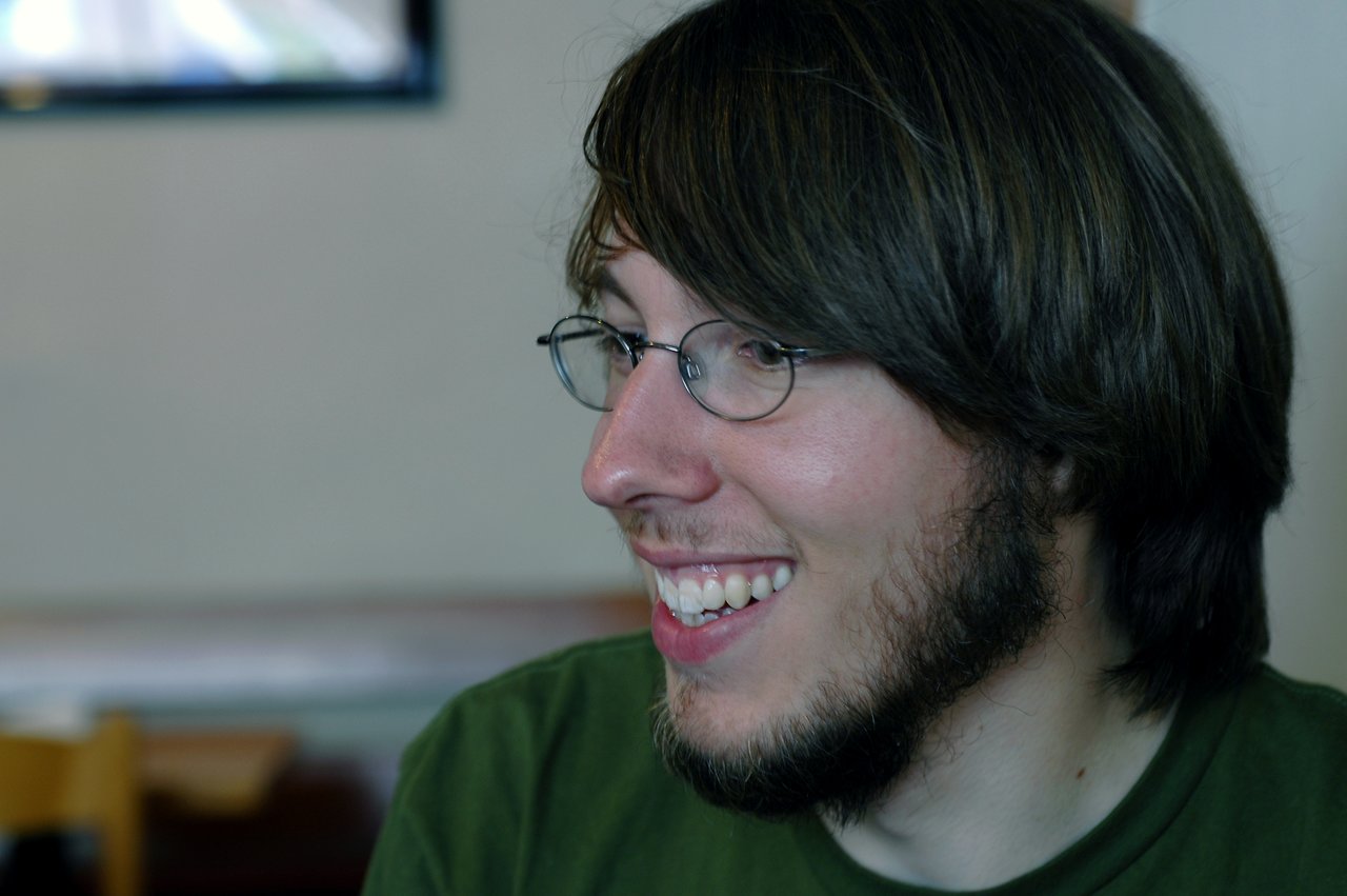 A man with glasses and a beard smiles while looking to the side in an indoor setting.