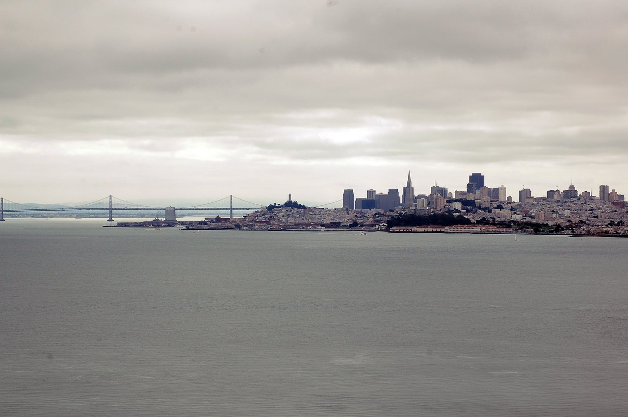 San Francisco skyline with the Golden Gate Bridge in the background, viewed across the water under a cloudy sky.
