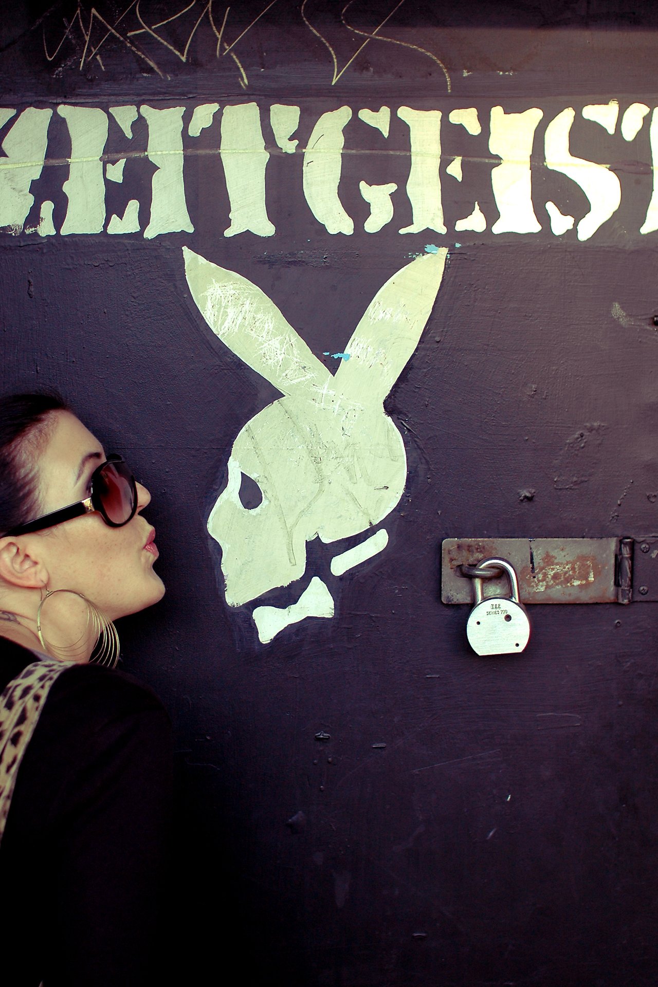 A woman wearing sunglasses playfully kisses a painted skull logo on a dark wall with "Zeitgeist" written above.