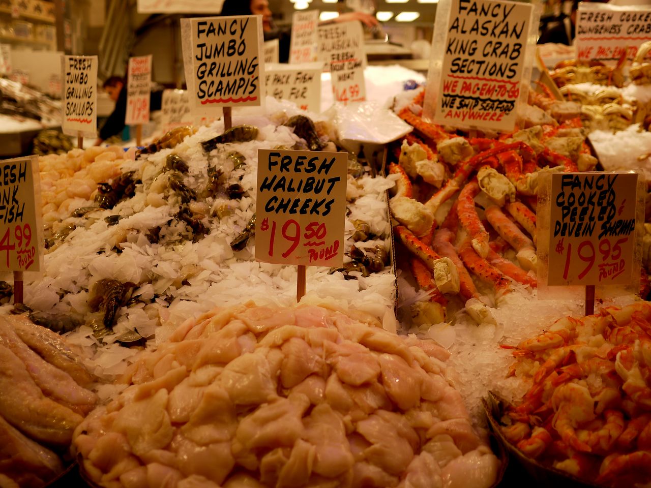 Fresh seafood display at a public market, featuring halibut cheeks, shrimp, scallops, and Alaskan king crab on ice.