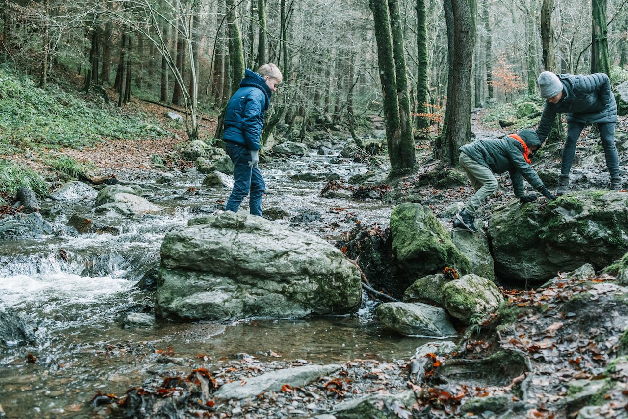 Three people hike through a rocky forest stream, with one helping another climb over a mossy boulder.