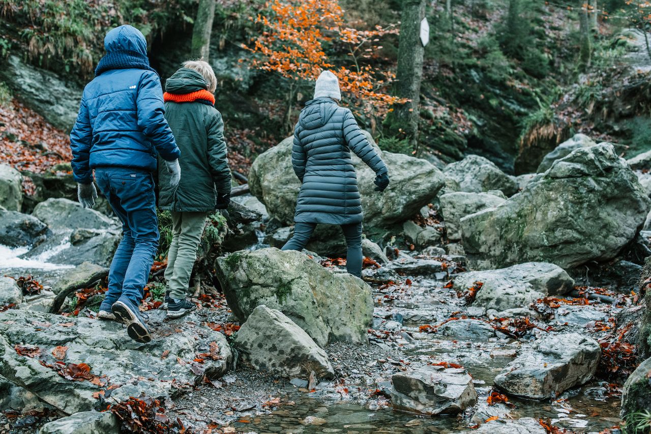 Three people in warm clothing hike over rocky terrain in a forested valley with a small stream.