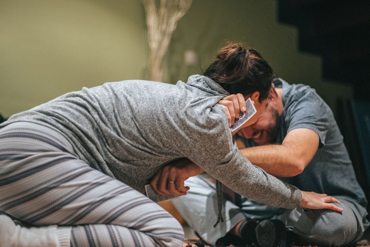 Two people laughing and playfully leaning on each other while holding playing cards in a casual indoor setting.