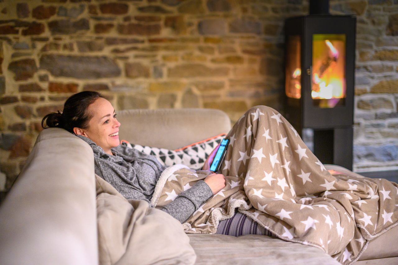 A woman relaxes on a couch under a blanket, smiling while looking at her phone near a fireplace.