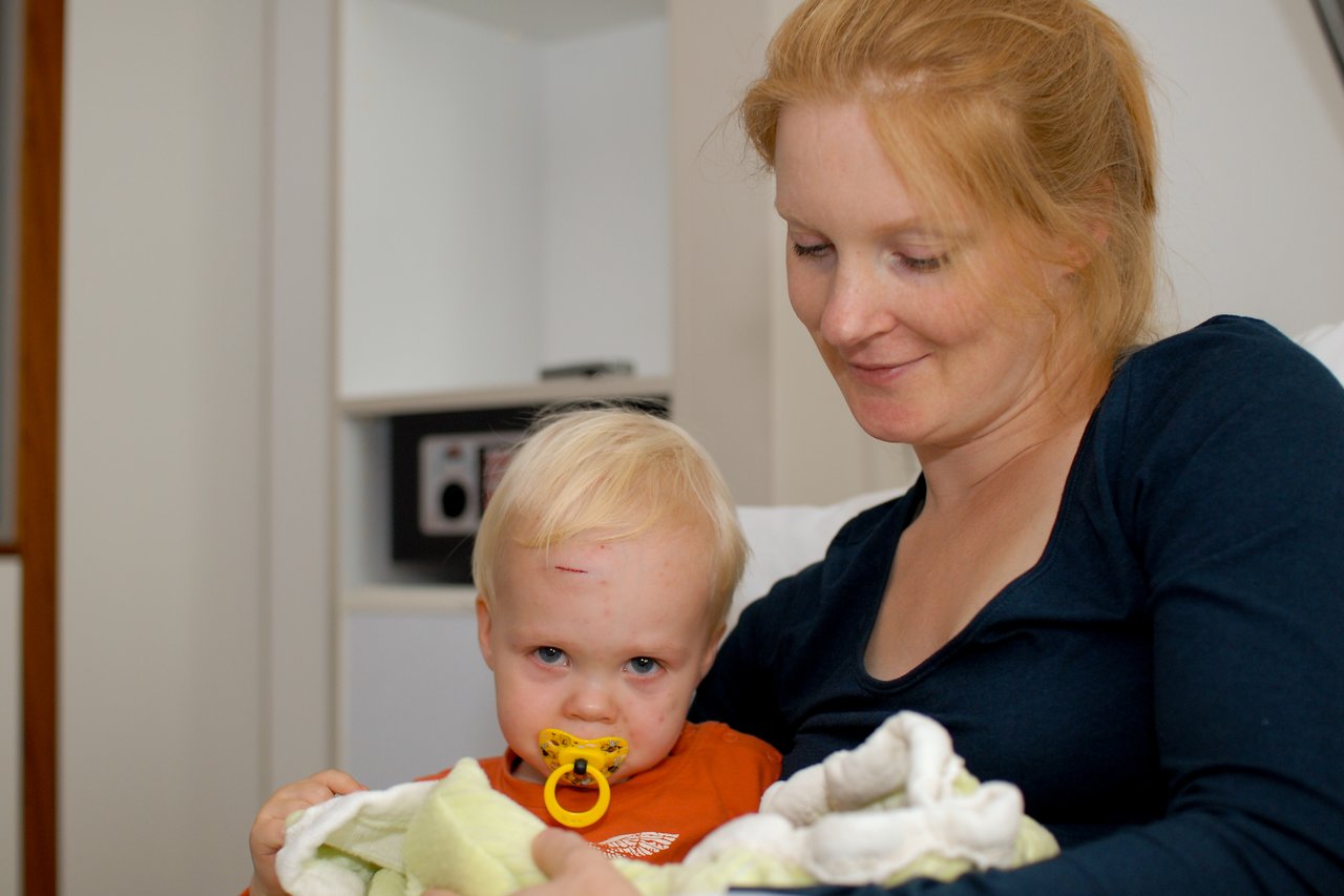 A woman holds a young child with a pacifier and a small forehead injury, both wrapped in blankets.