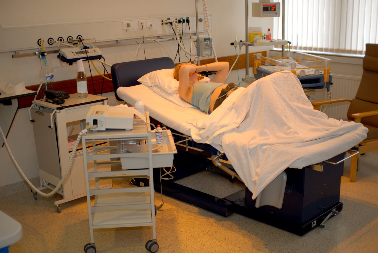 A woman lies in a hospital bed in a delivery room, with medical equipment nearby.