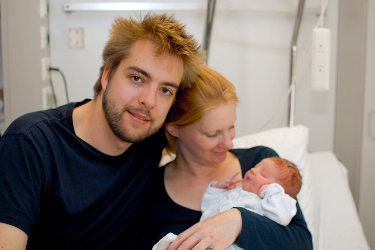 A man and woman sit together in a hospital room, with the woman holding a newborn baby in her arms.