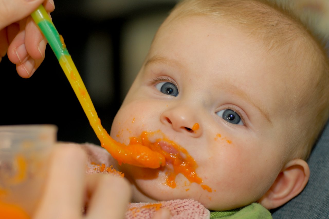 A baby with food on their face is being fed orange vegetable puree from a spoon.