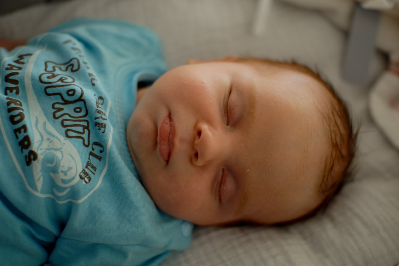 A newborn baby sleeps on a soft surface, wearing a blue shirt with printed text and graphics.