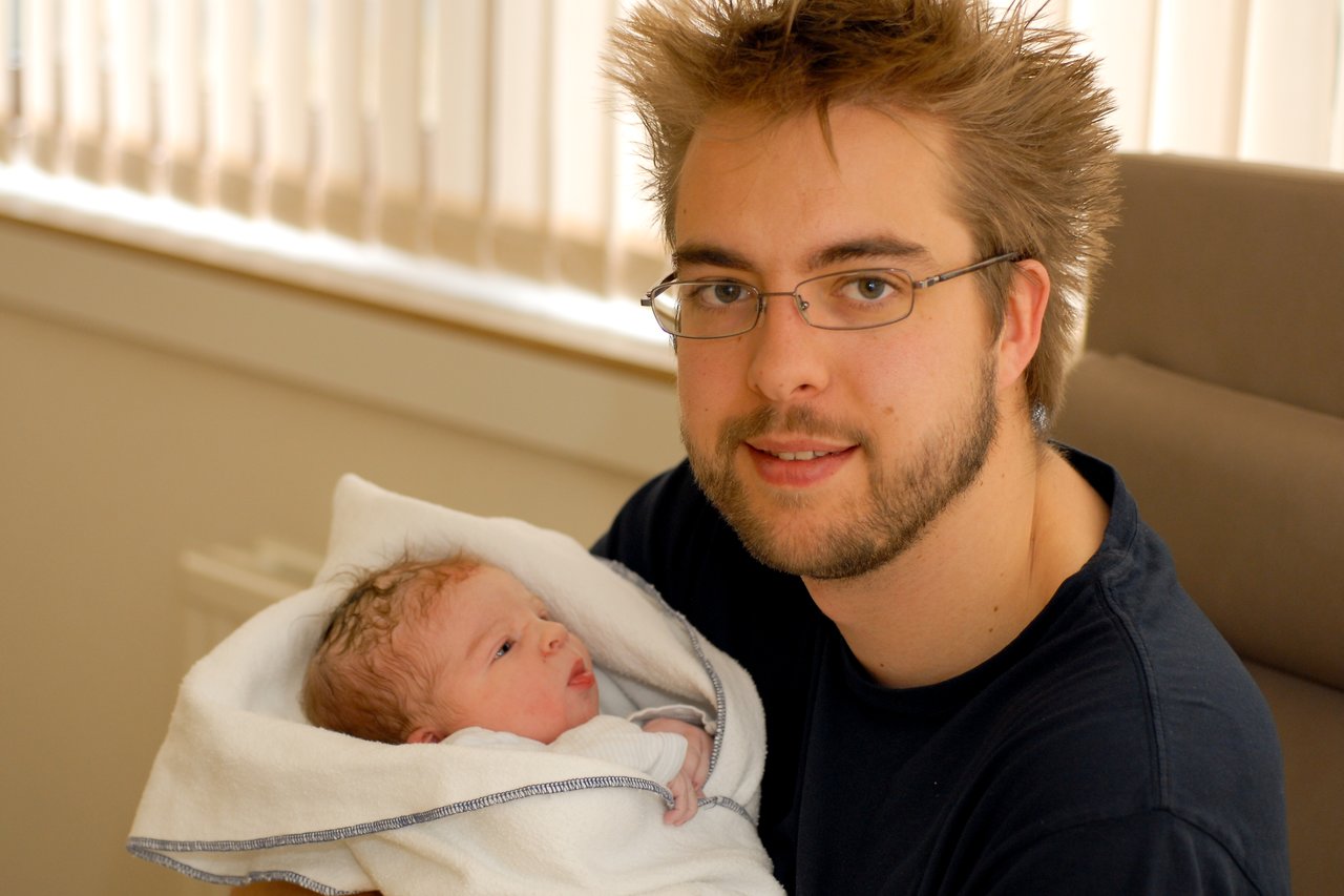 A man with glasses and a beard holds a newborn baby wrapped in a white towel, looking at the camera.