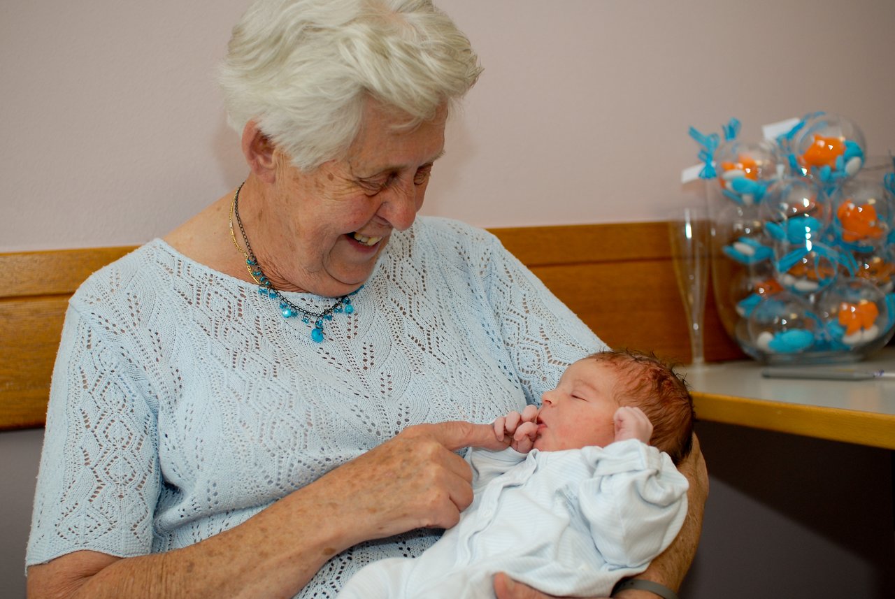 An elderly woman smiles while holding a newborn baby, who grasps her finger.