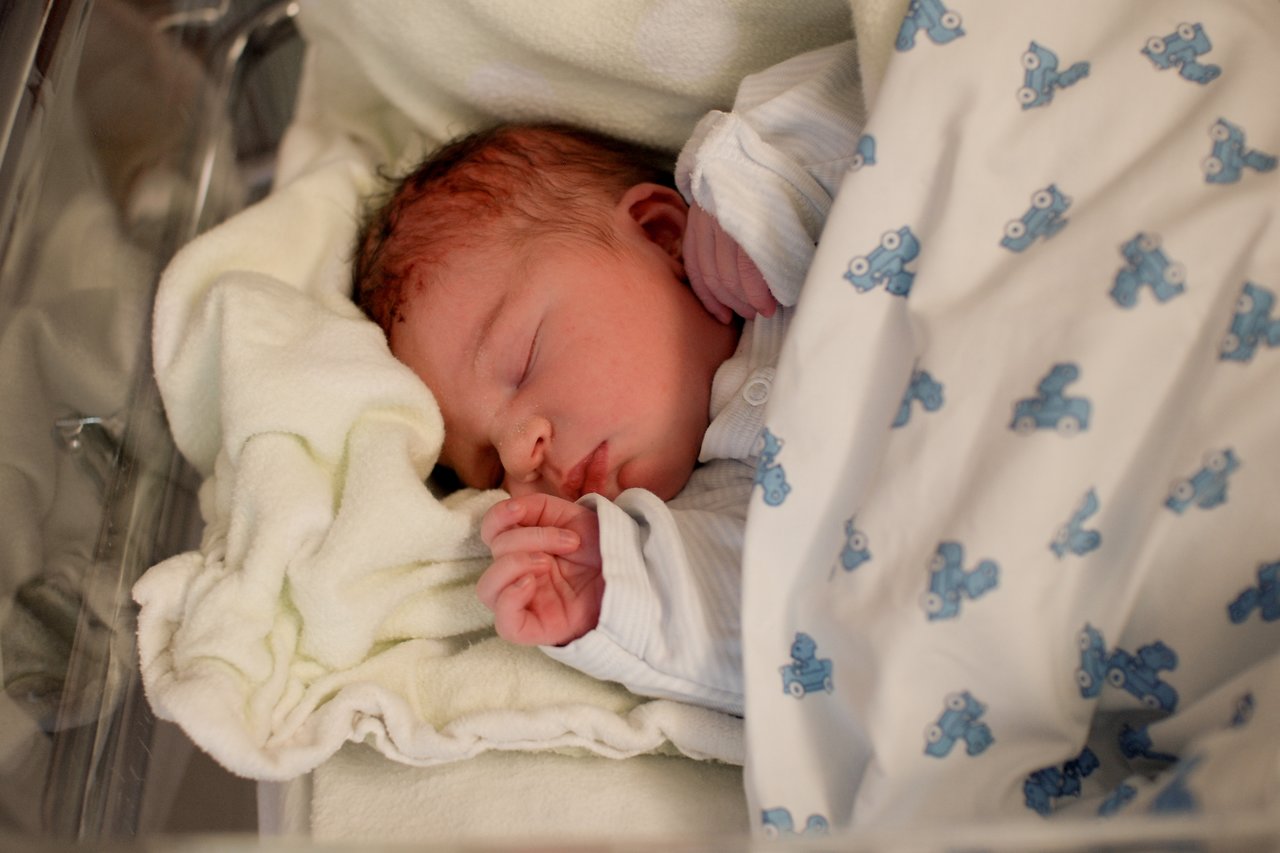A newborn baby sleeps peacefully, wrapped in a blanket with blue car patterns, resting on soft bedding.