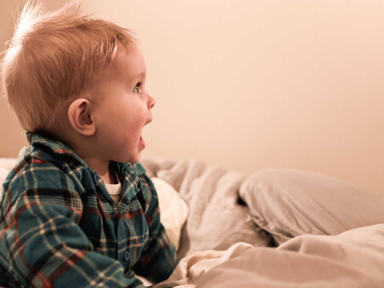 A baby in plaid pajamas sits on a bed, looking excited and surprised.