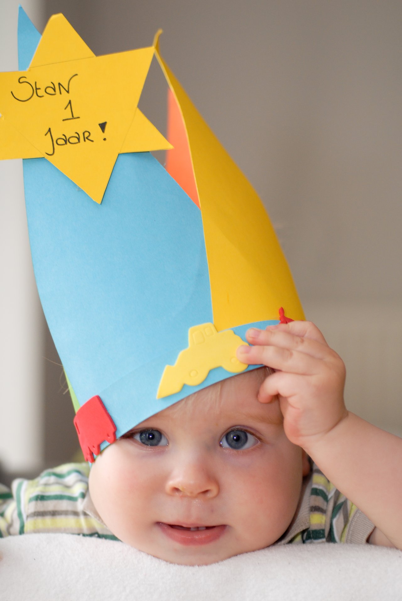A baby wearing a colorful paper hat with "Stan 1 Jaar!" written on it, celebrating their first birthday.