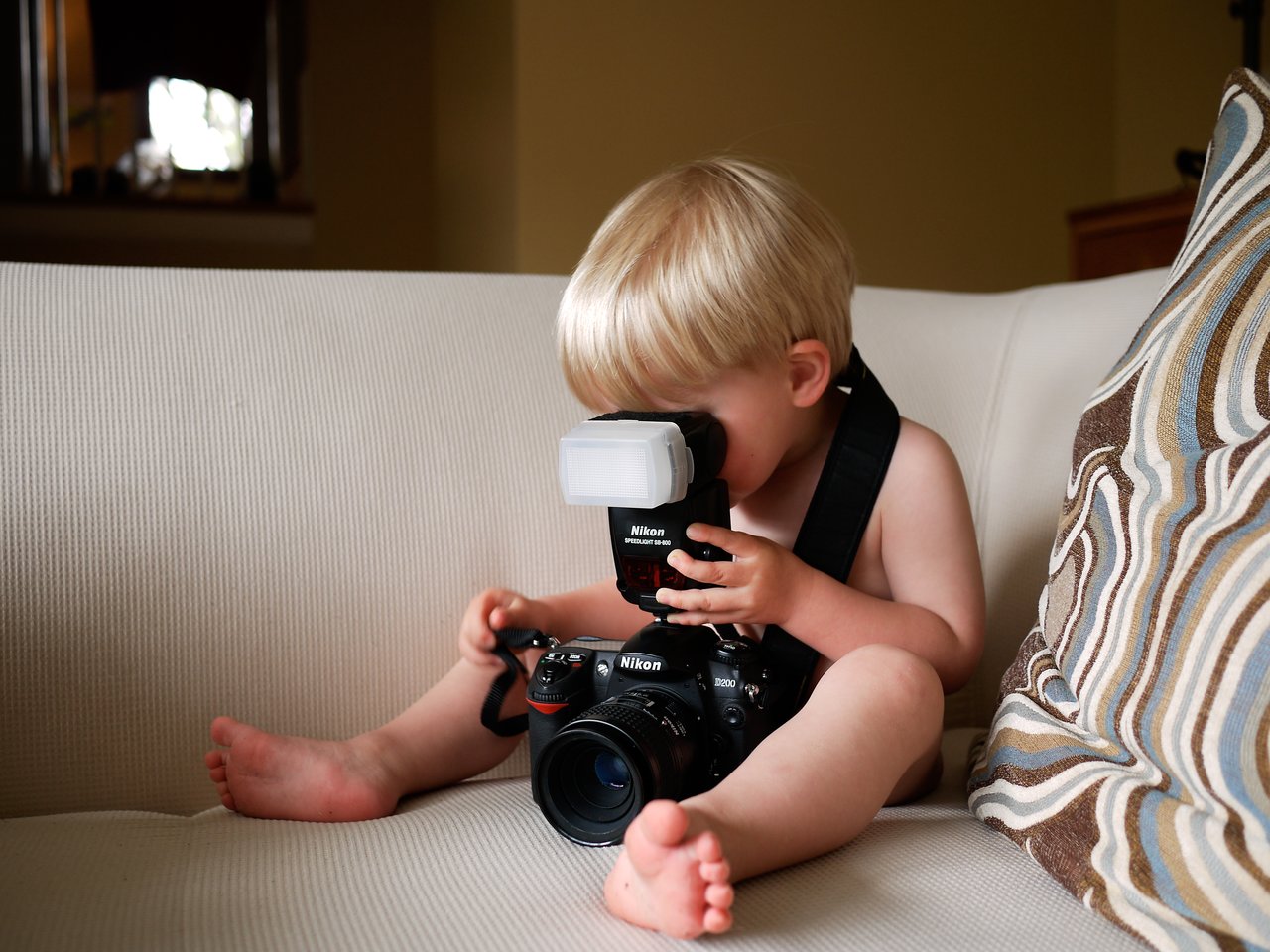 A young child sits on a couch, holding a large camera and looking through the viewfinder.