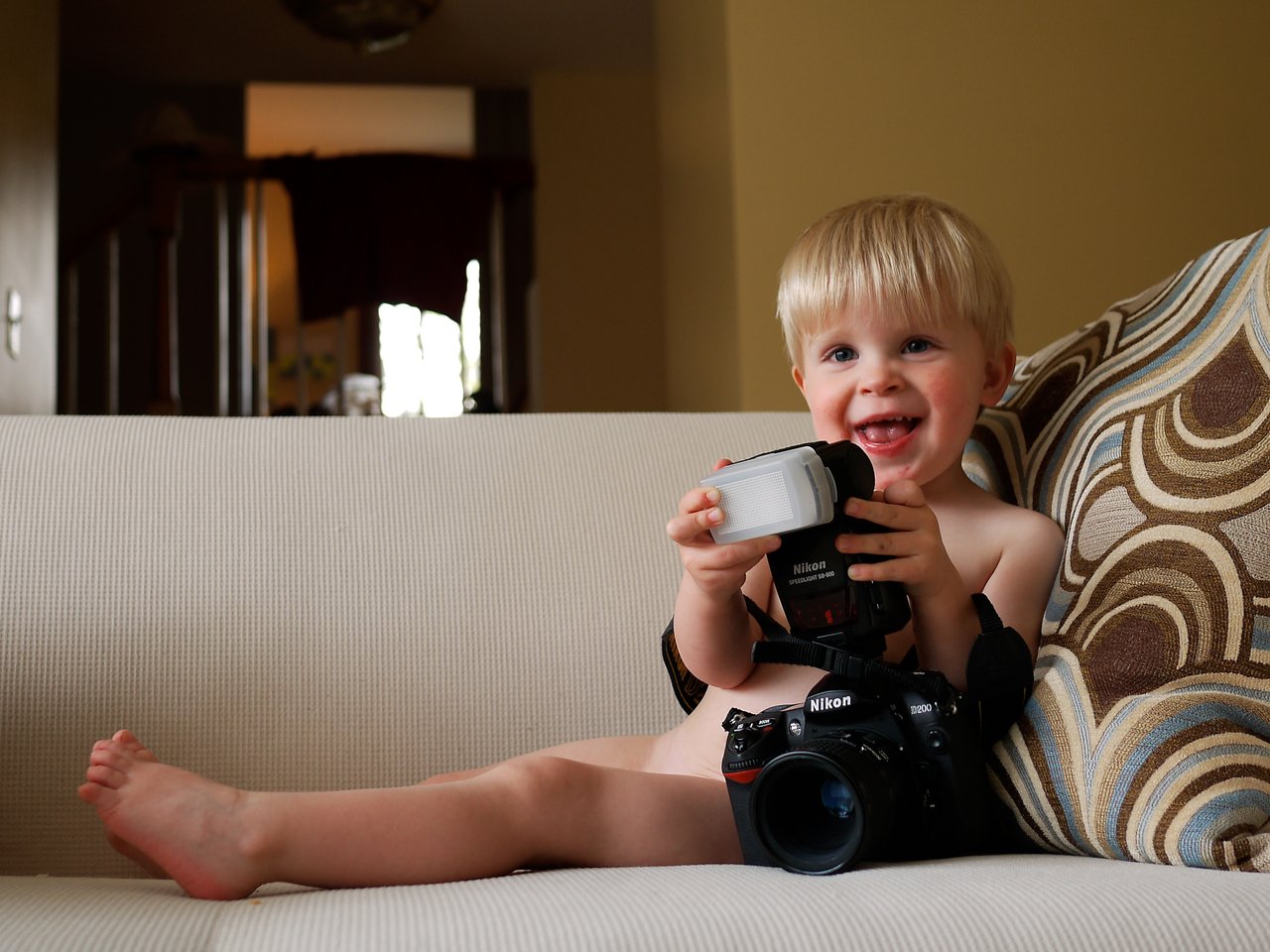 A smiling child sits on a couch holding a camera flash, with a Nikon camera resting on their lap.