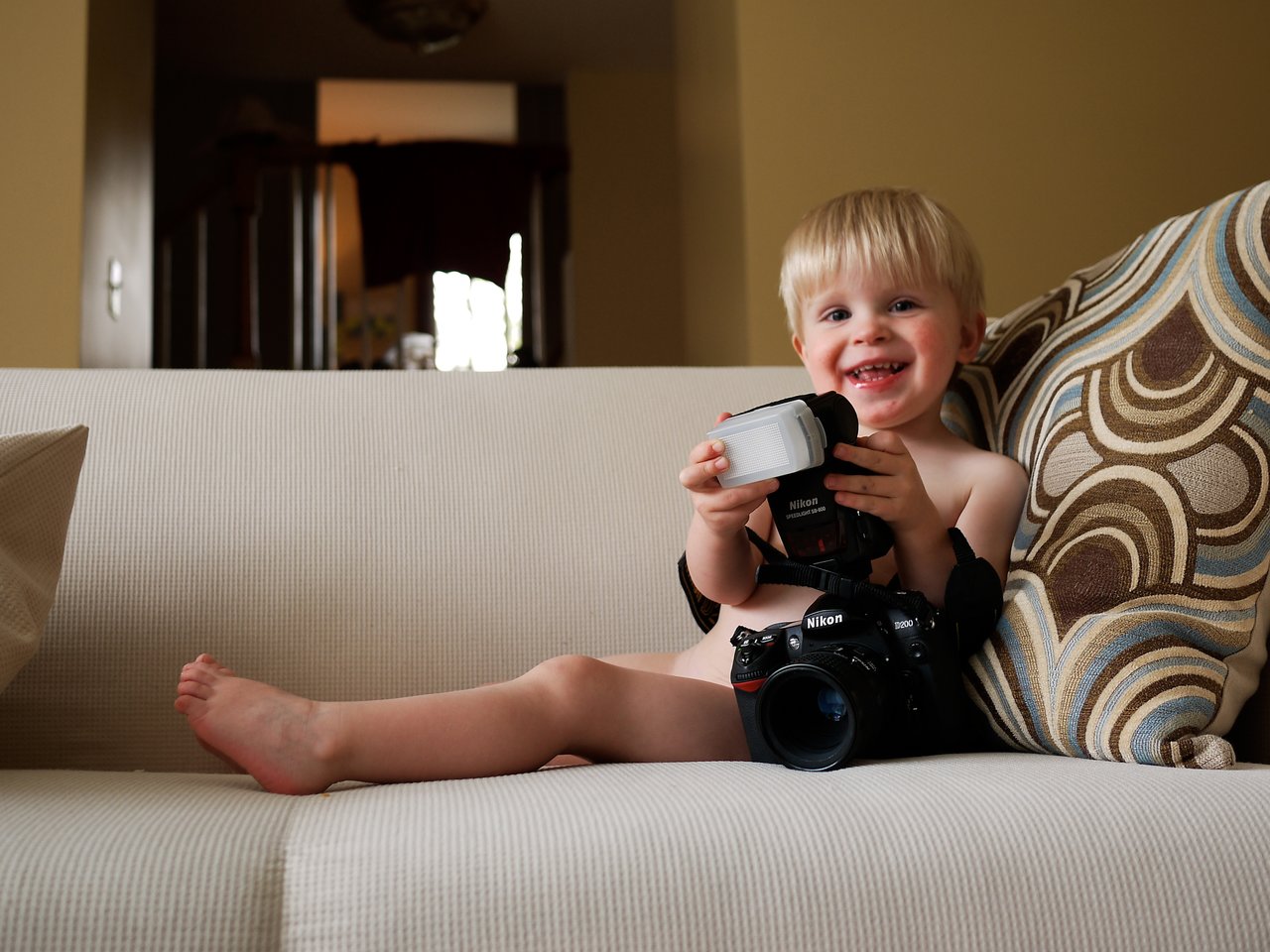 A smiling child sits on a couch holding a camera with a flash attachment.
