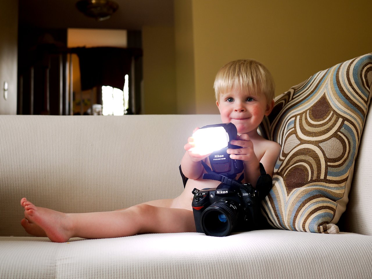 A young child sits on a couch holding a camera with a bright flash, looking curious and engaged.