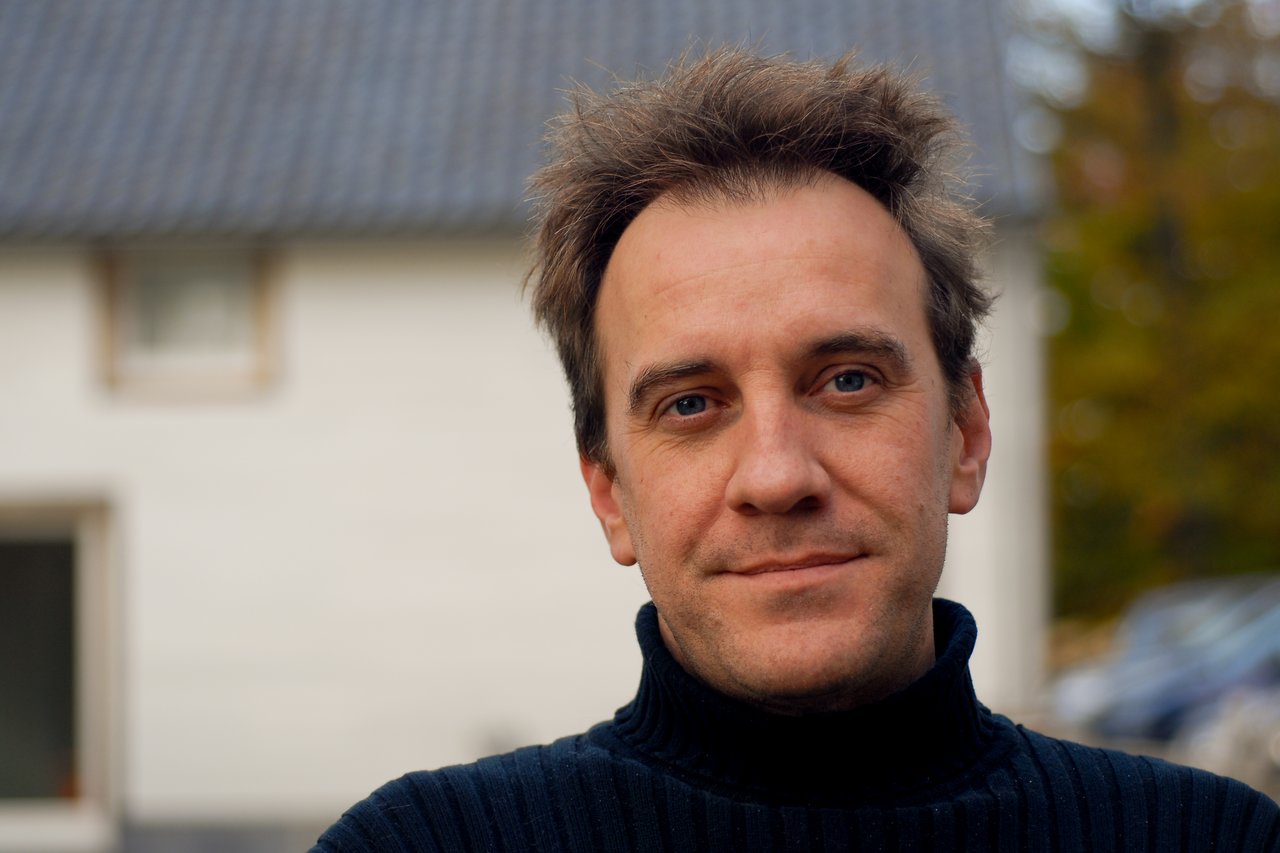 A man with short, slightly messy hair wears a black turtleneck sweater and looks directly at the camera, smiling slightly.