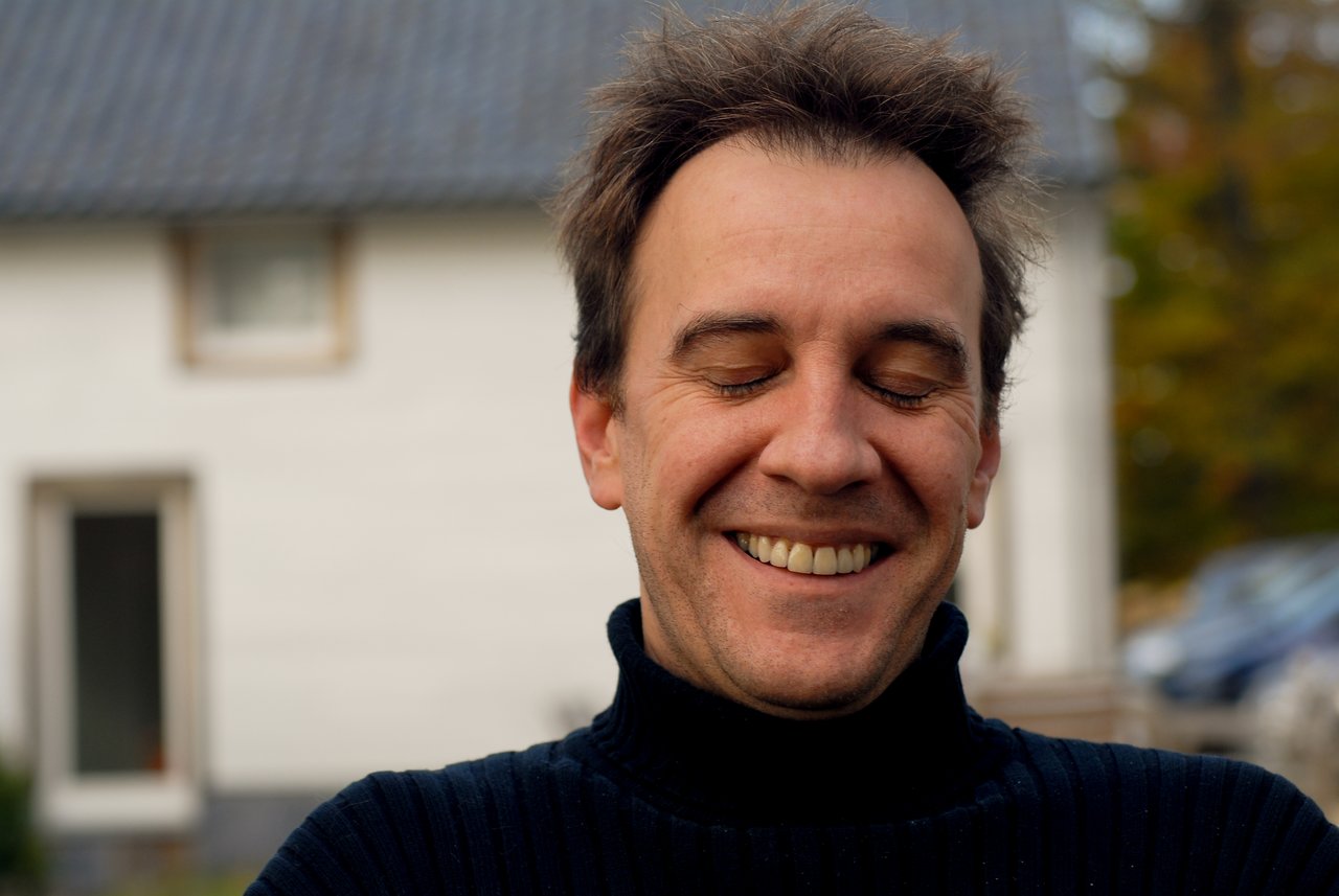 A man with short, messy hair smiles with his eyes closed, wearing a black turtleneck sweater outdoors.