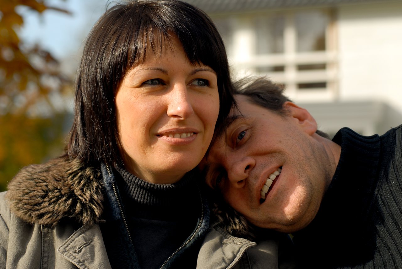A man leans his head on a woman's shoulder while both look in different directions, smiling slightly.