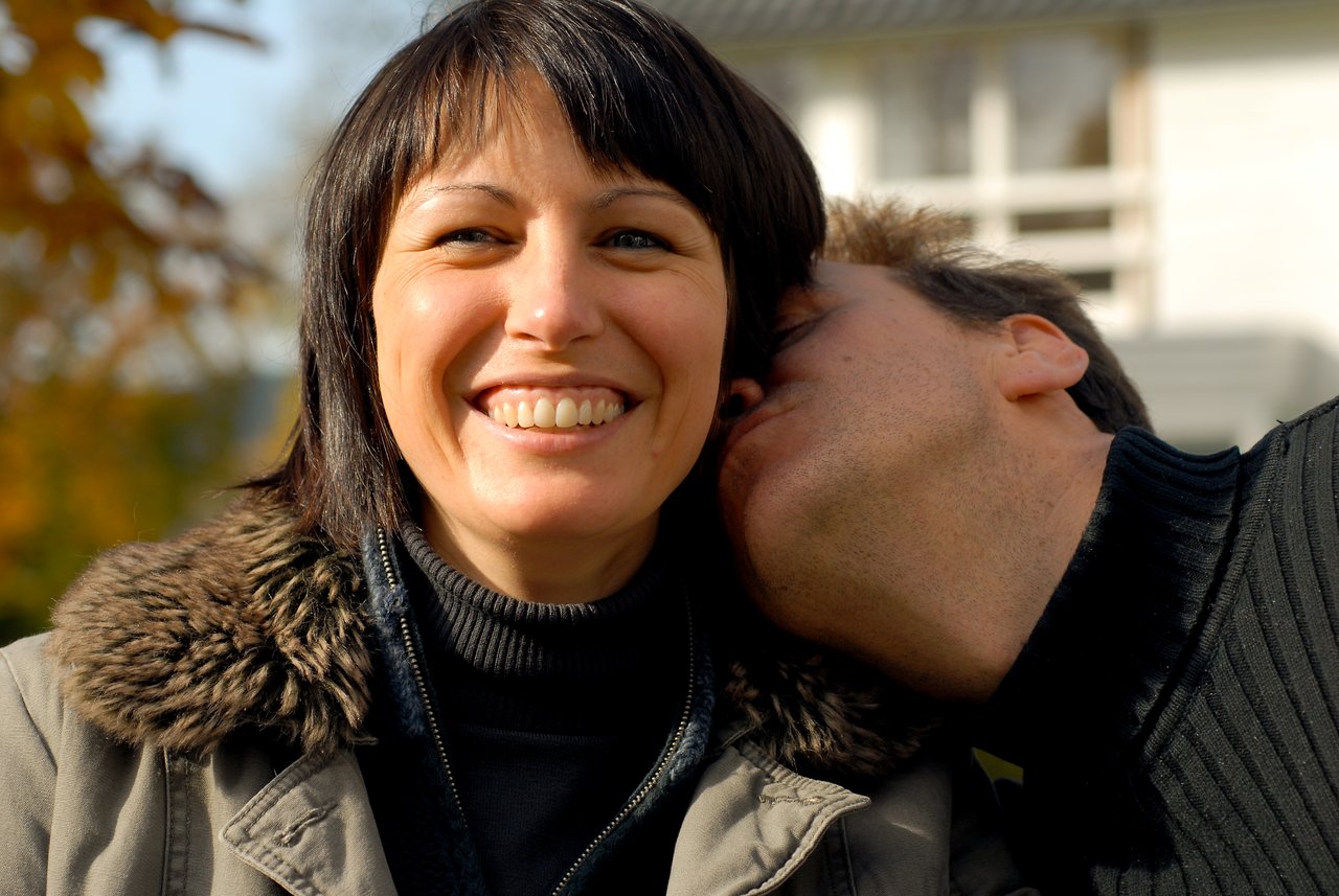 A smiling woman in a winter coat as a man leans in to kiss her on the cheek.