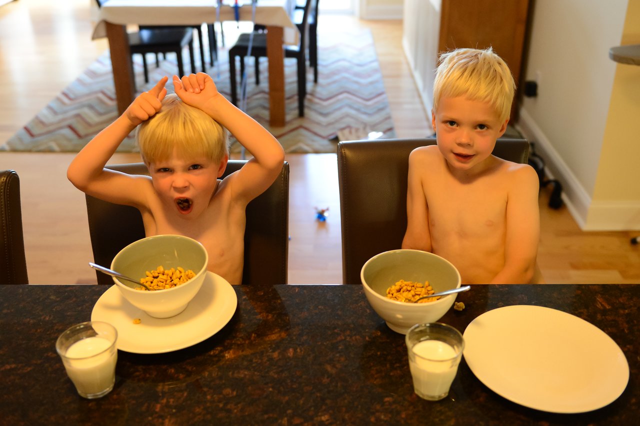 Two young boys sit at a table with cereal and milk; one is making a playful gesture, the other is calm.