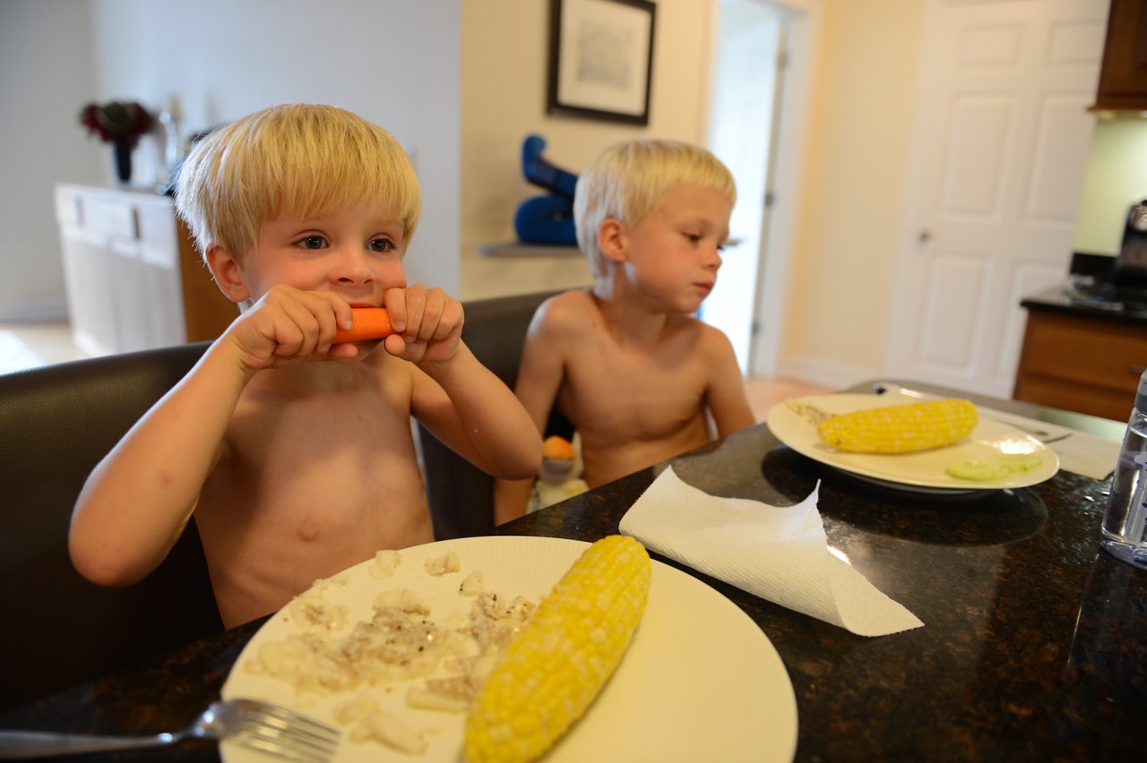 Two young boys sit at a kitchen table, one eating a carrot while the other looks away.